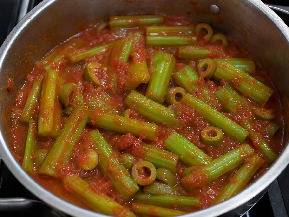 Celery pieces and halved green olives in a thick tomato sauce inside a wide braising pot
