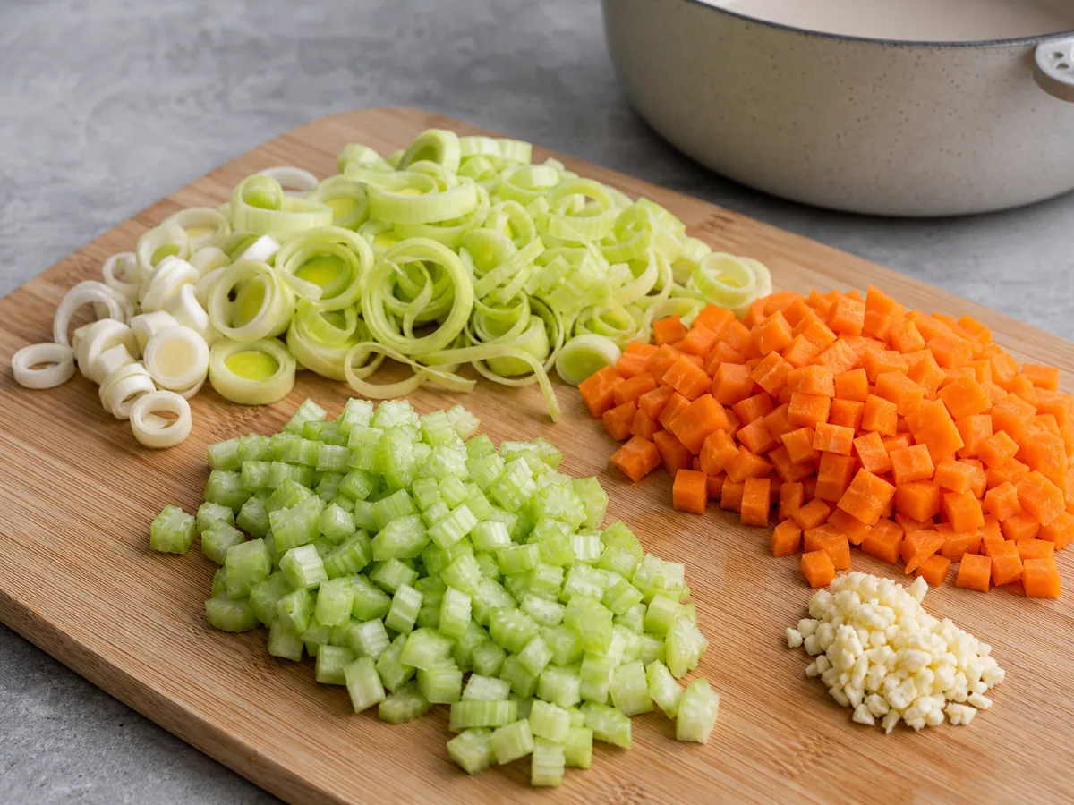Cleaned leeks sliced crosswise beside evenly cut carrots, celery, and minced garlic