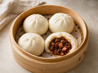 Vaporera de bambú llena de bao de char siu esponjosos, con uno abierto para mostrar el relleno de cerdo
