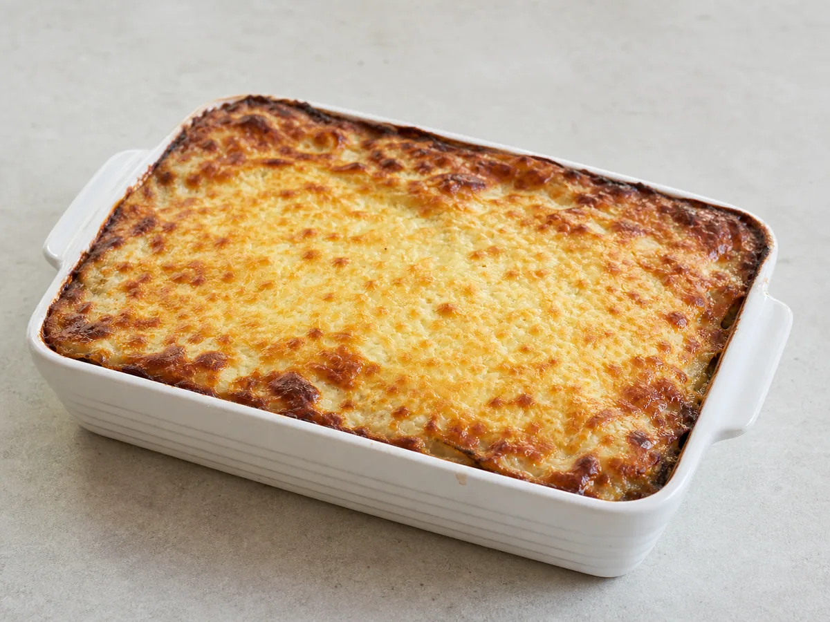 Moussaka griega de cordero horneada y muy dorada, con bordes burbujeantes y un termómetro reviseo el centro caliente