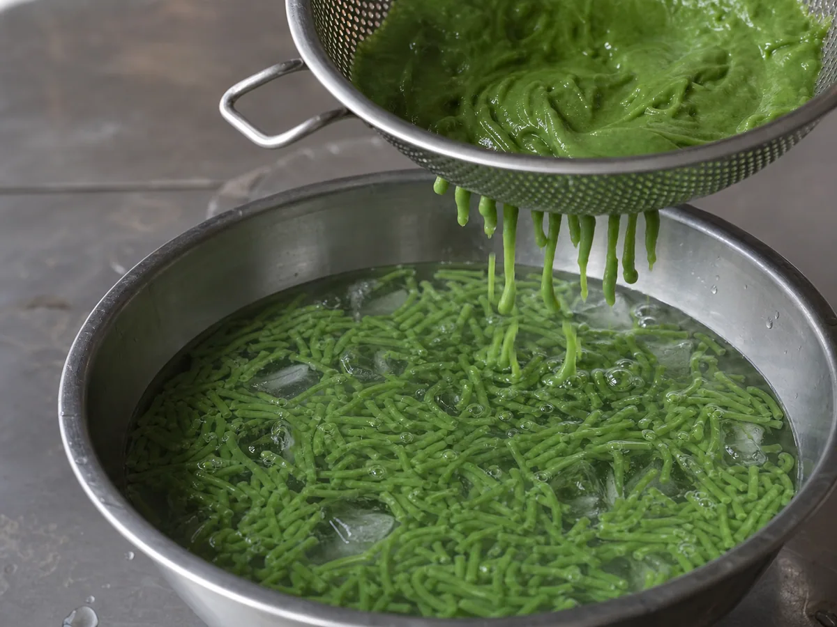 Tiras verdes de cendol recién prensadas que se afirman bajo una prensa de cendol en agua con hielo