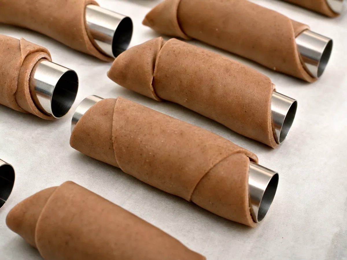 Masa fina de cannoli teñida con cacao, envuelta y sellada alrededor de tubos metálicos antes de freír