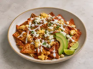 Chilaquiles rojos cubiertos con huevos revueltos, queso fresco, crema, aguacate, cebolla y cilantro