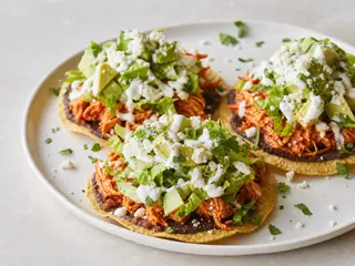Dos tostadas de tinga de pollo con frijoles negros, lechuga, aguacate, Cotija, crema y cilantro