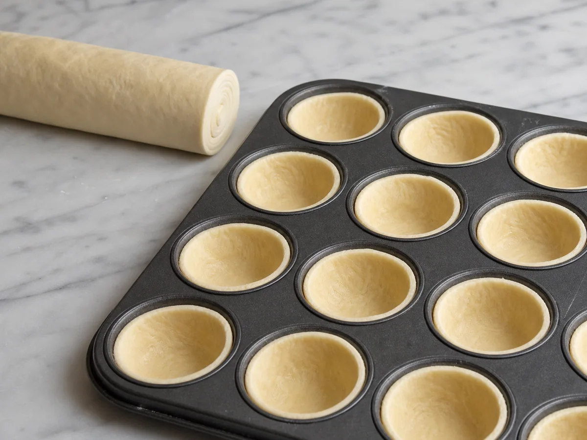 Puff pastry rounds being pressed into thin shells inside a metal muffin tin