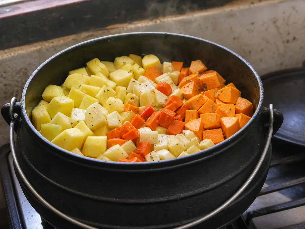 Potjie de frango totalmente cozido, com camadas distintas de raízes macias e um termômetro verificando uma sobrecoxa exposta