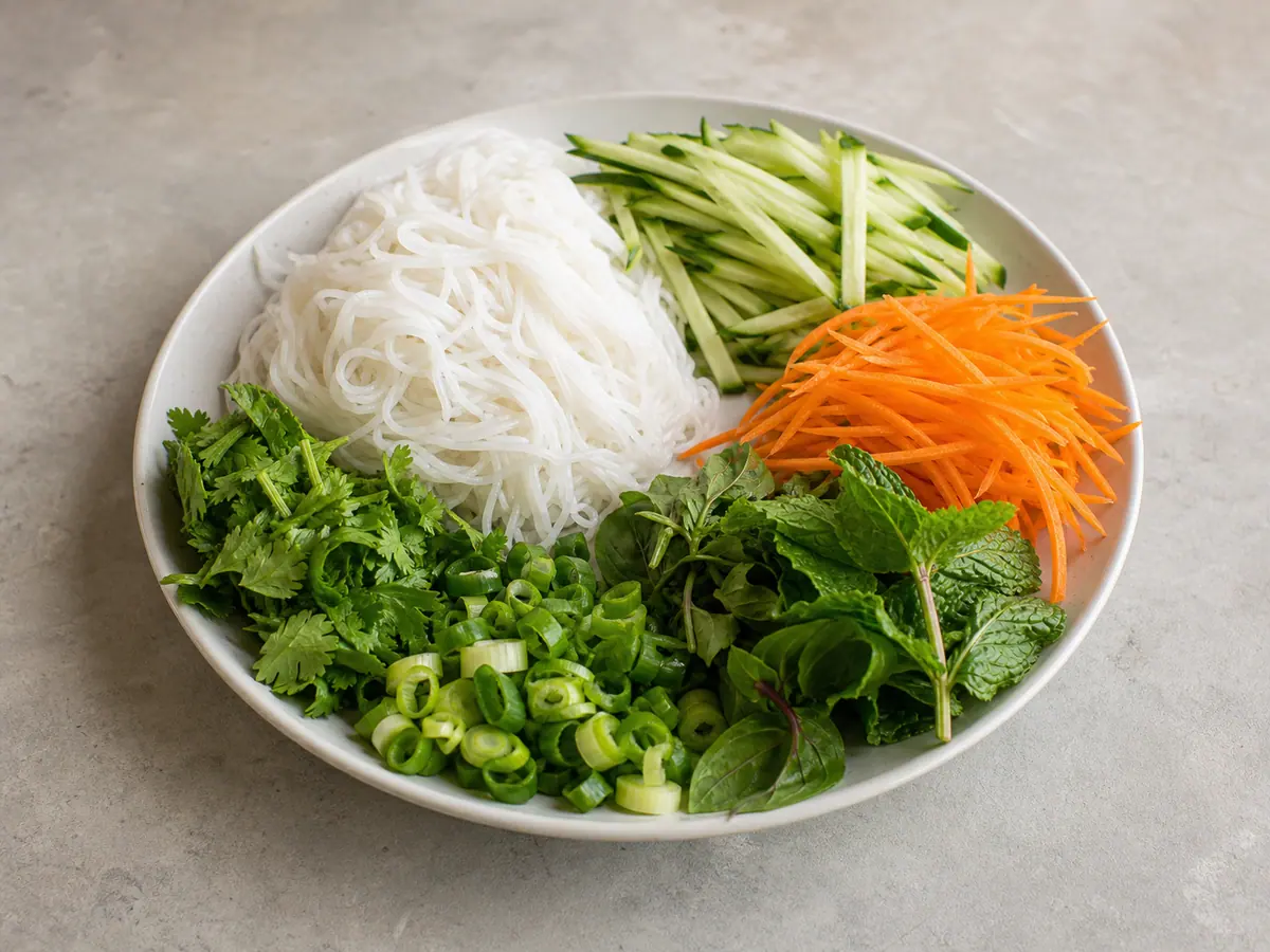 Drained springy rice vermicelli beside prepared vegetables, herbs, and peanuts